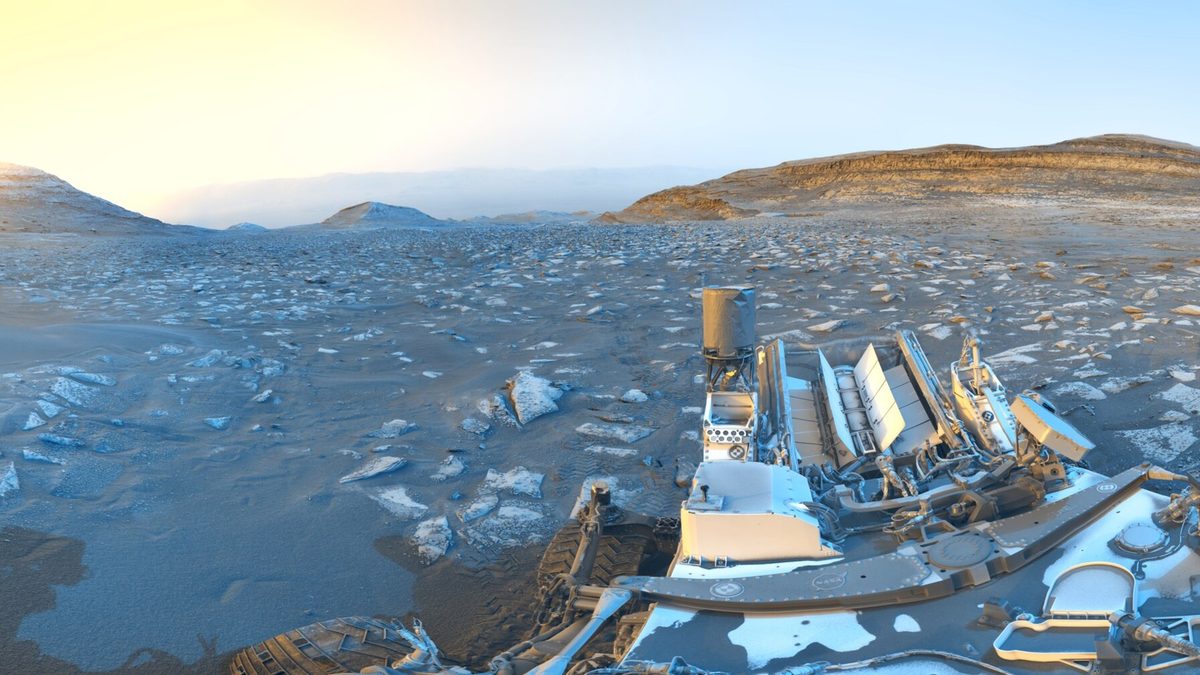 Parte do jipe-robô Curiosity sobre solo rochoso de Marte, com morros ao fundo, em tons de azul e amarelo