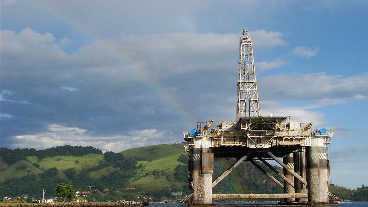 Plataforma de petróleo perto da costa, com morros verdes e arco-íris ao fundo
