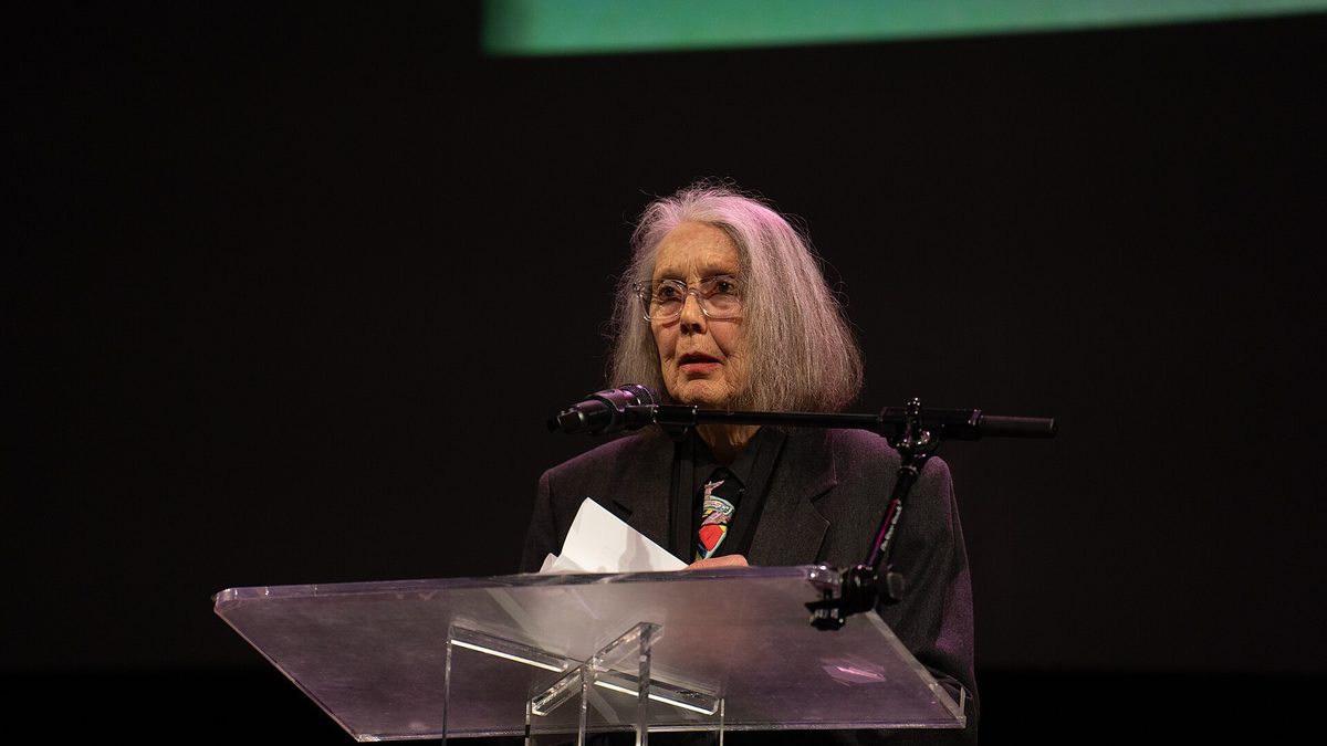 A poeta Anne Carson lê ao microfone, diante de um púlpito transparente, em palco escuro