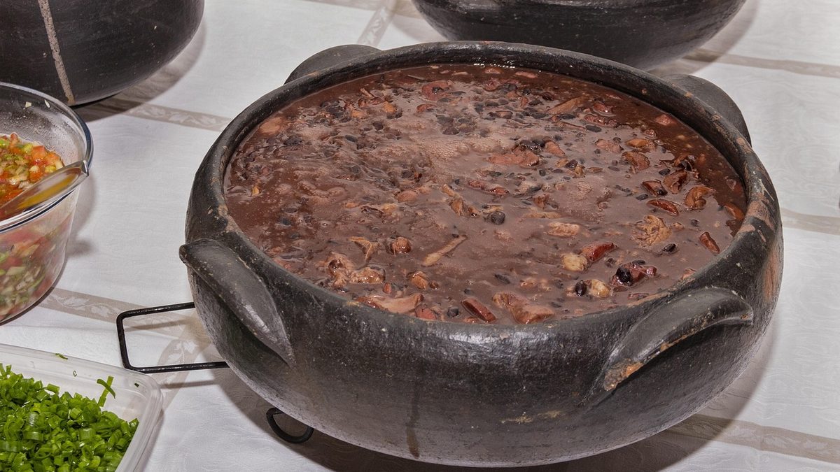 Feijoada em panela de barro sobre bufê, ao lado de saladas