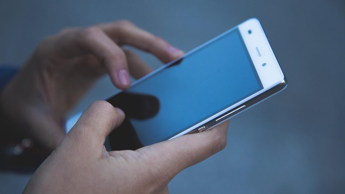 Mãos seguram um celular branco inclinado, diante de fundo cinza desfocado