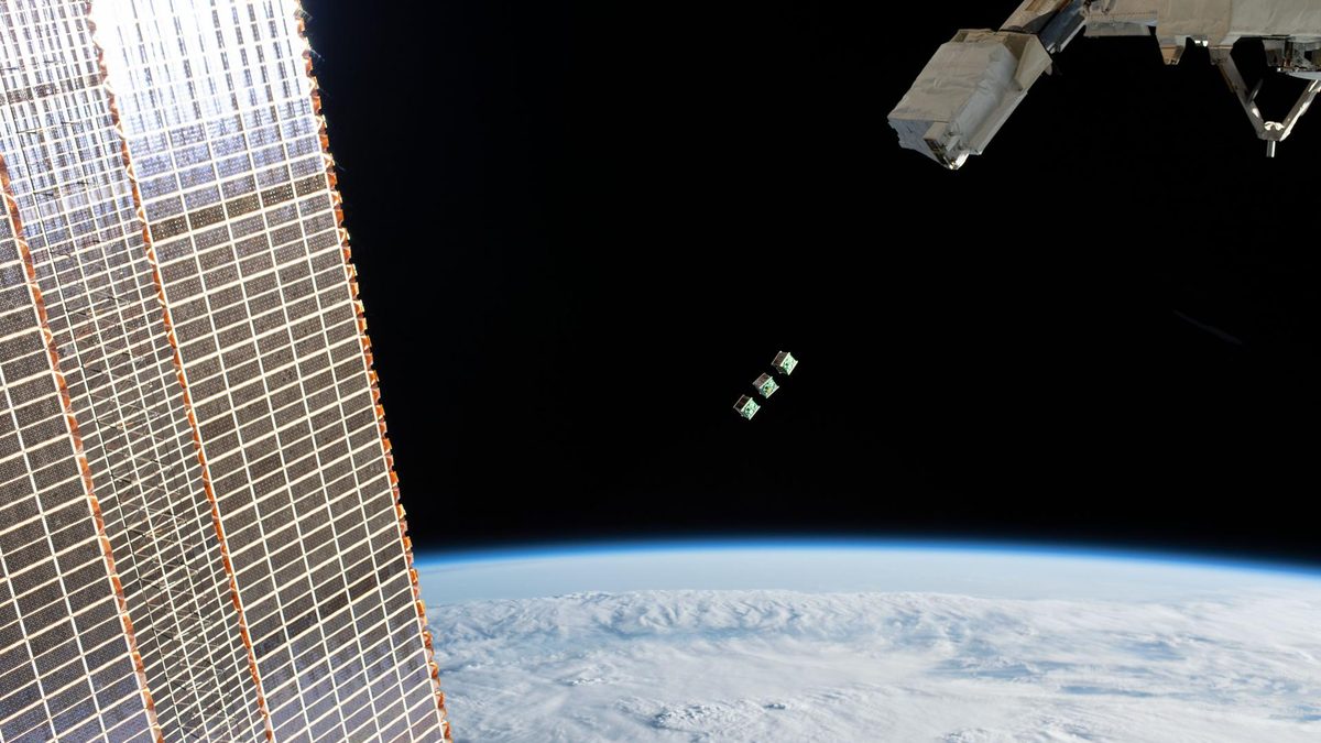 Três pequenos satélites no espaço, entre o painel solar da estação espacial e a Terra coberta de nuvens
