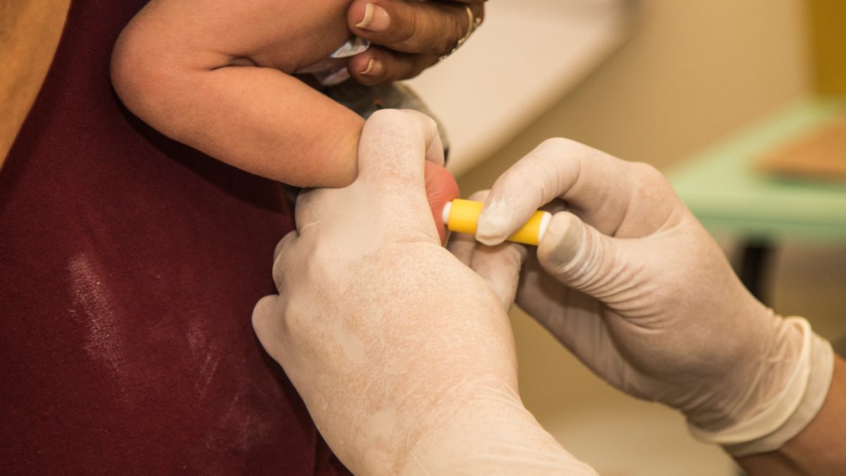 Mãos com luvas furam o calcanhar de um bebê com lanceta para o teste do pezinho
